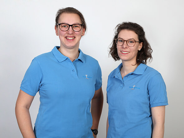 Zwei Frauen in blauen Poloshirts posieren lächelnd vor einem neutralen Hintergrund.
