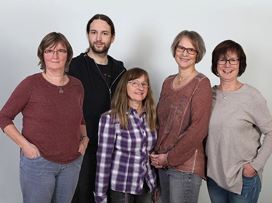 Gruppe von fünf Personen, die lächelnd vor einem neutralen Hintergrund posieren.