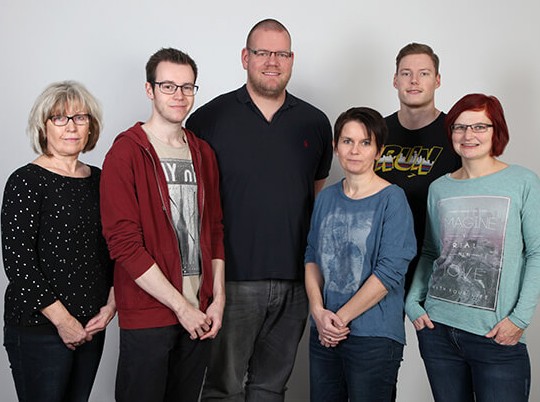 Sechs Personen posieren lächelnd vor einem neutralen Hintergrund.
