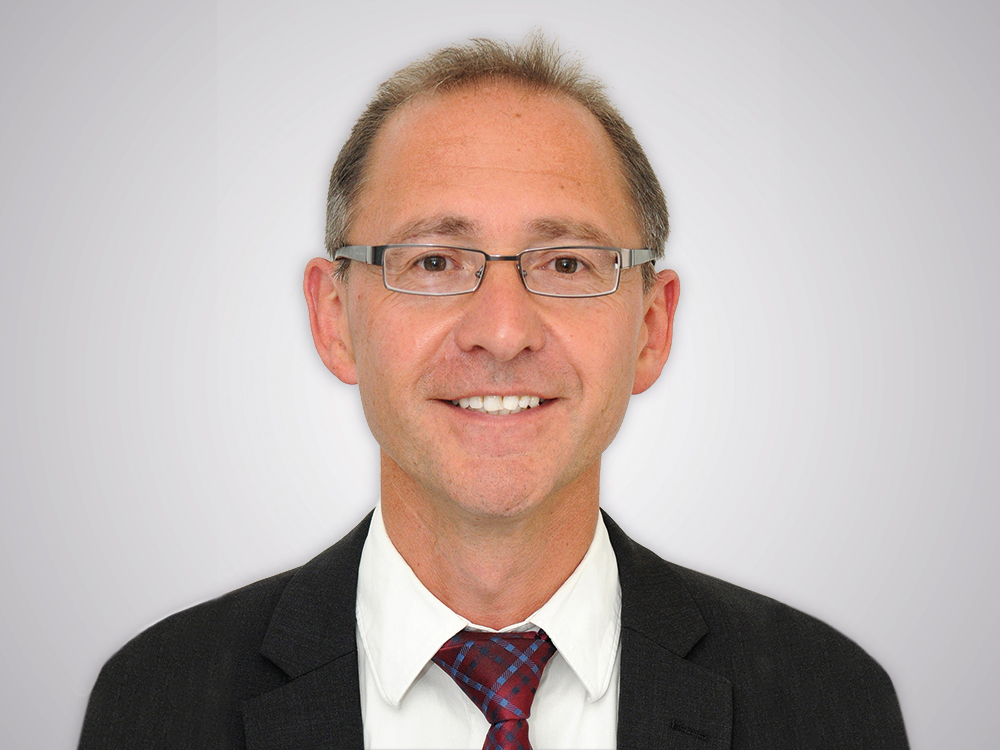 Mann mit Brille, lächelt freundlich, trägt Anzug und Krawatte, neutrale Hintergrundgestaltung.
