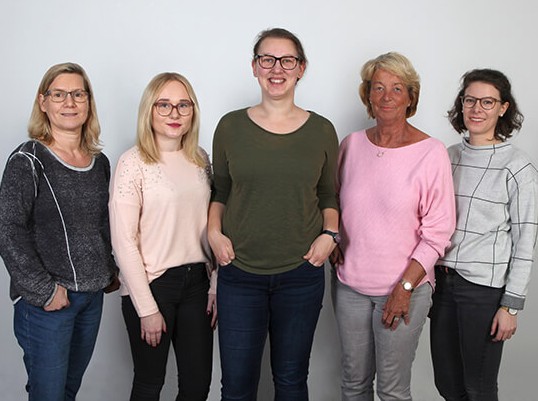 Fünf Frauen stehen vor neutralem Hintergrund in modischer Kleidung.