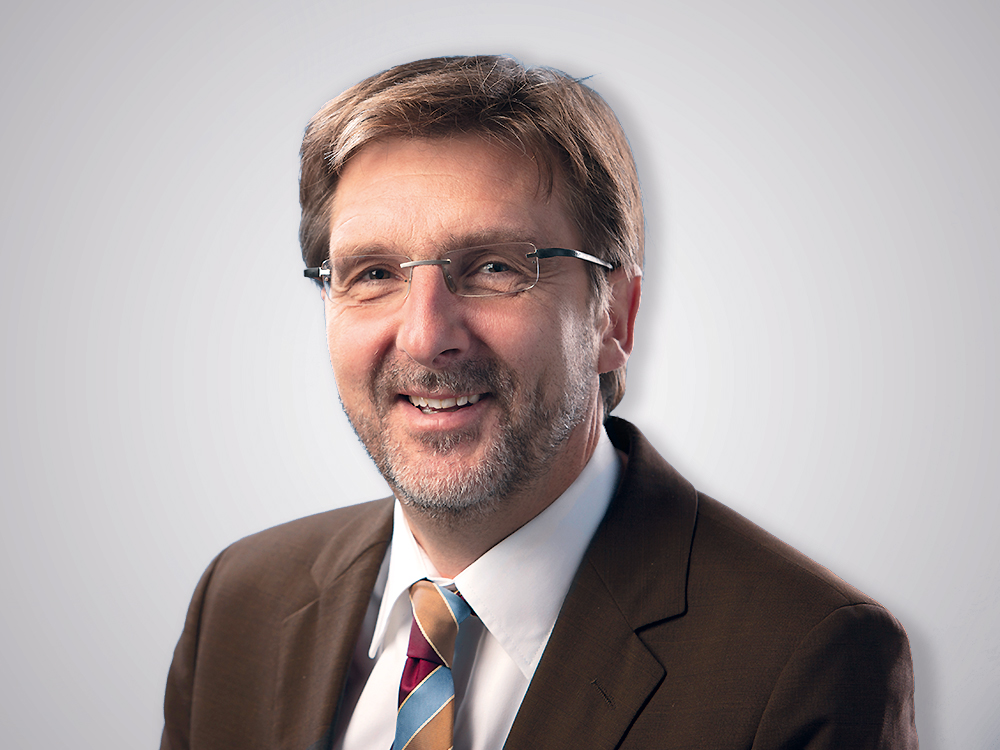 Mann in Brille und Anzug lächelt freundlich vor einem hellen Hintergrund.